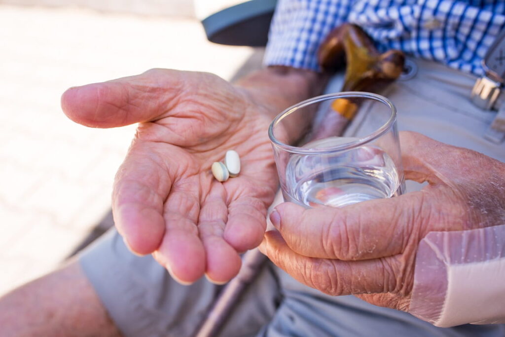 Hände eines älteren Mannes halten Wasserglas und Tabletten