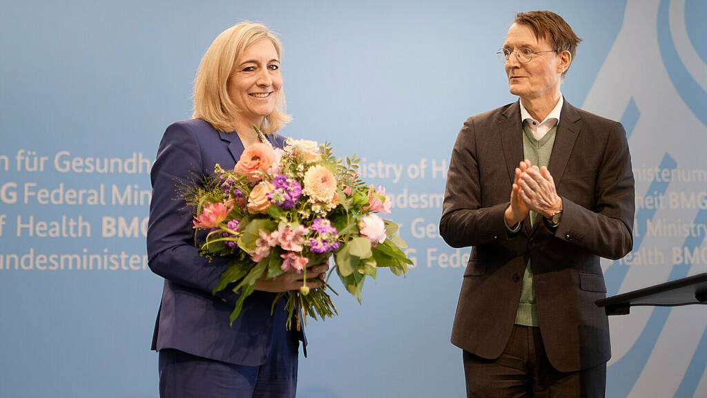 Offizielle Veranstaltung im Bundesministerium für Gesundheit mit Blumenübergabe vor blauem Hintergrund mit mehrsprachigem Ministeriumslogo.