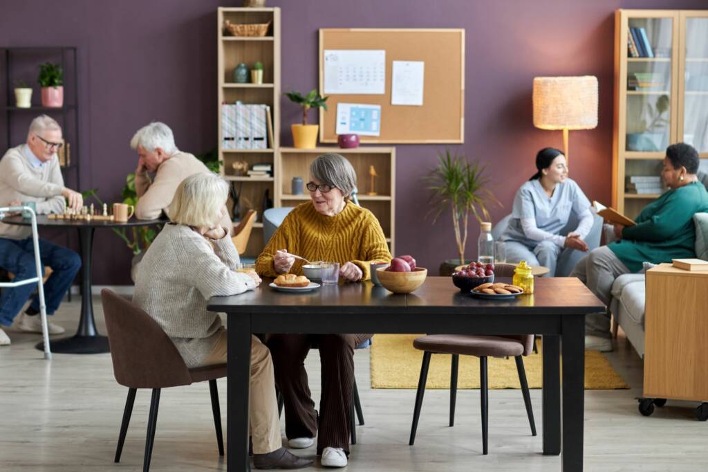 Gemeinschaftsraum mit Senioren beim Essen, Schachspielen und Gesprächen in gemütlicher Wohnatmosphäre mit Pflanzen und Pinnwand.