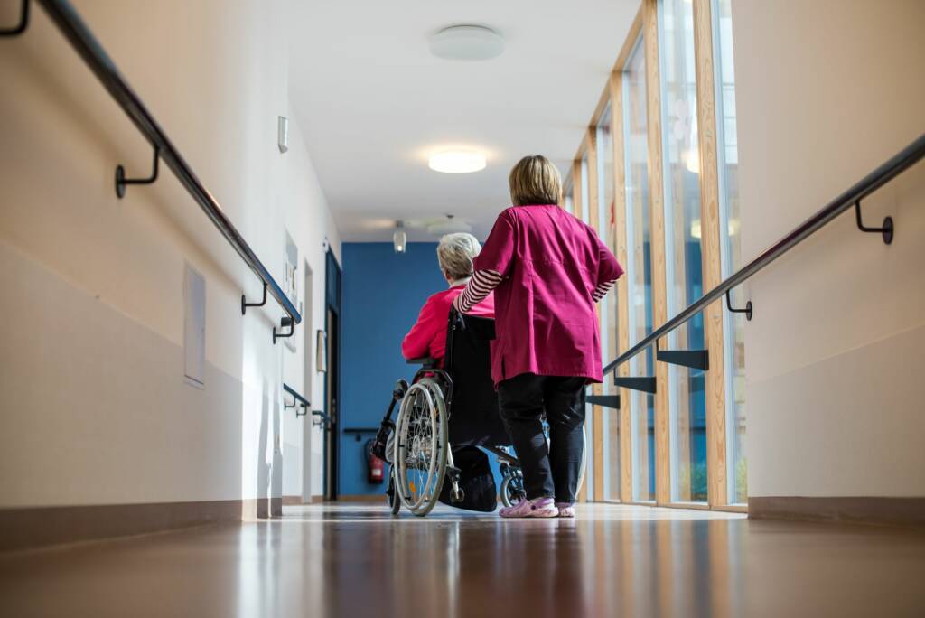 Frau schiebt Patientin im Rollstuhl über einen Flur im Krankenhaus oder Pflegeheim.