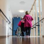Frau schiebt Patientin im Rollstuhl über einen Flur im Krankenhaus oder Pflegeheim.