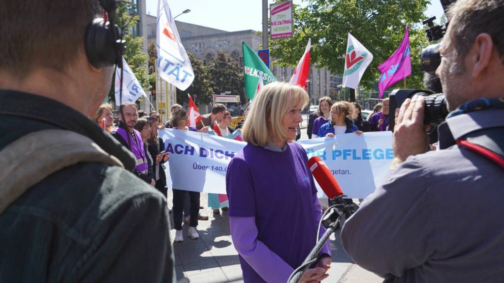 Demonstranten in lila Kleidung bei Pflegeprotest, eine Person gibt Interview, Banner und Gewerkschaftsflaggen im Hintergrund.