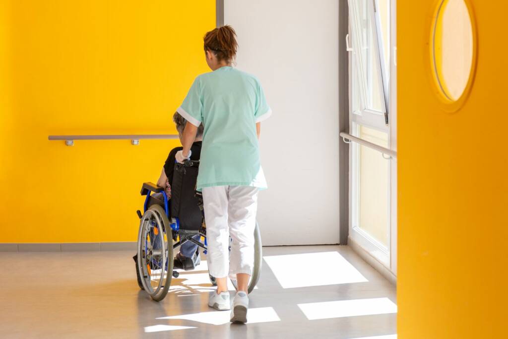 flegekraft in mint-grüner Kleidung schiebt Rollstuhl durch modernen Krankenhausflur mit gelber Wand.
