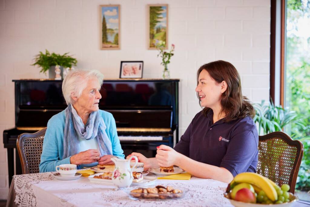 Betreuungskraft und ältere Dame beim gemeinsamen Tee und Gebäck im Wohnzimmer mit Klavier und Pflanzen im Hintergrund.