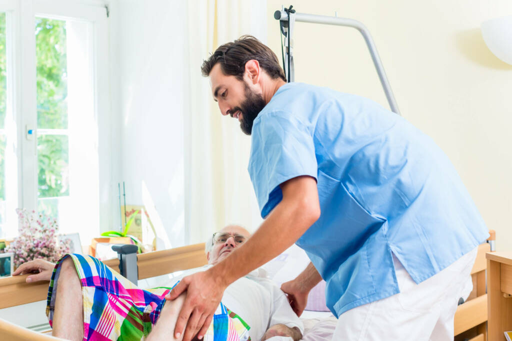 Elderly care nurse helping senior man from wheelchair to bed