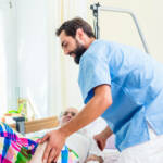 Elderly care nurse helping senior man from wheelchair to bed